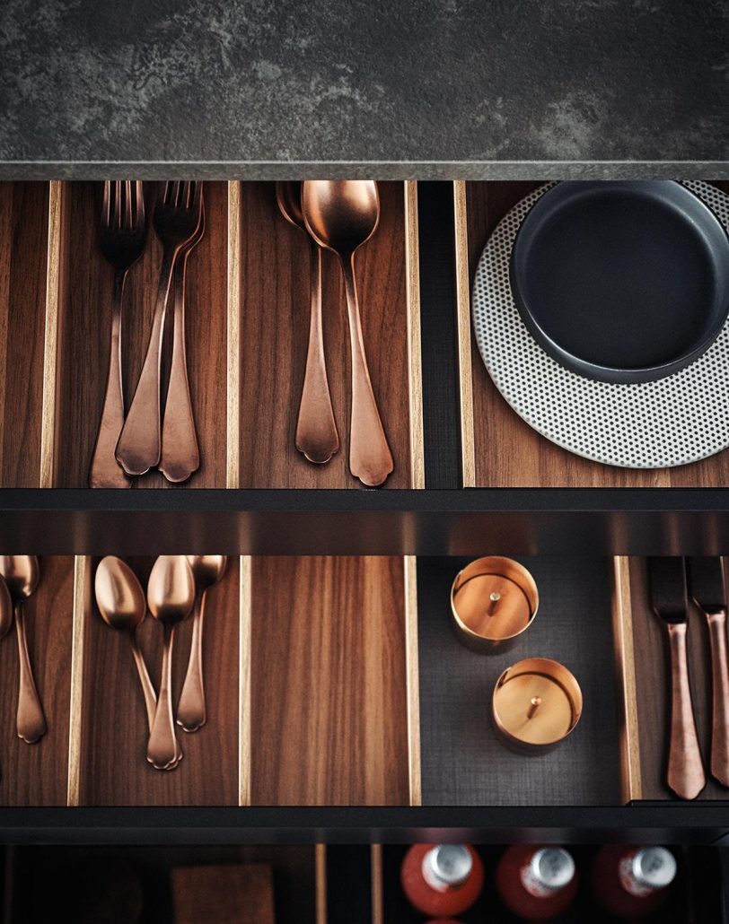 Detailansicht einer Küchenschublade in warmem Nussholz mit kupferfarbenem Besteck und matt-schwarzer Steinarbeitsplatte.