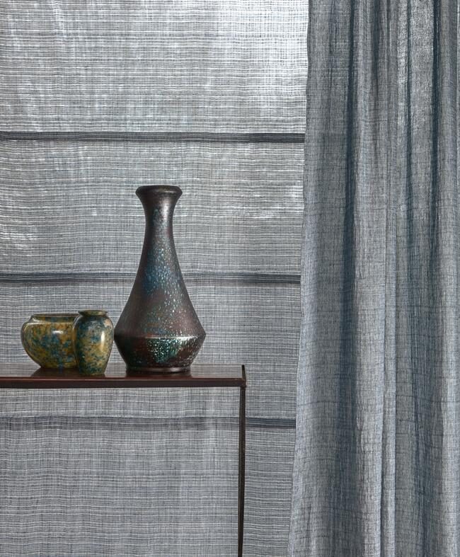 Stillleben mit schimmernder Keramikvase und silbrigem Leinenvorhang in gedämpftem Tageslicht.